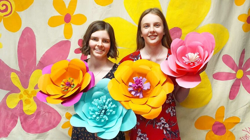 Two girls holding large paper flowers in front of a background painted with yellow flowers.