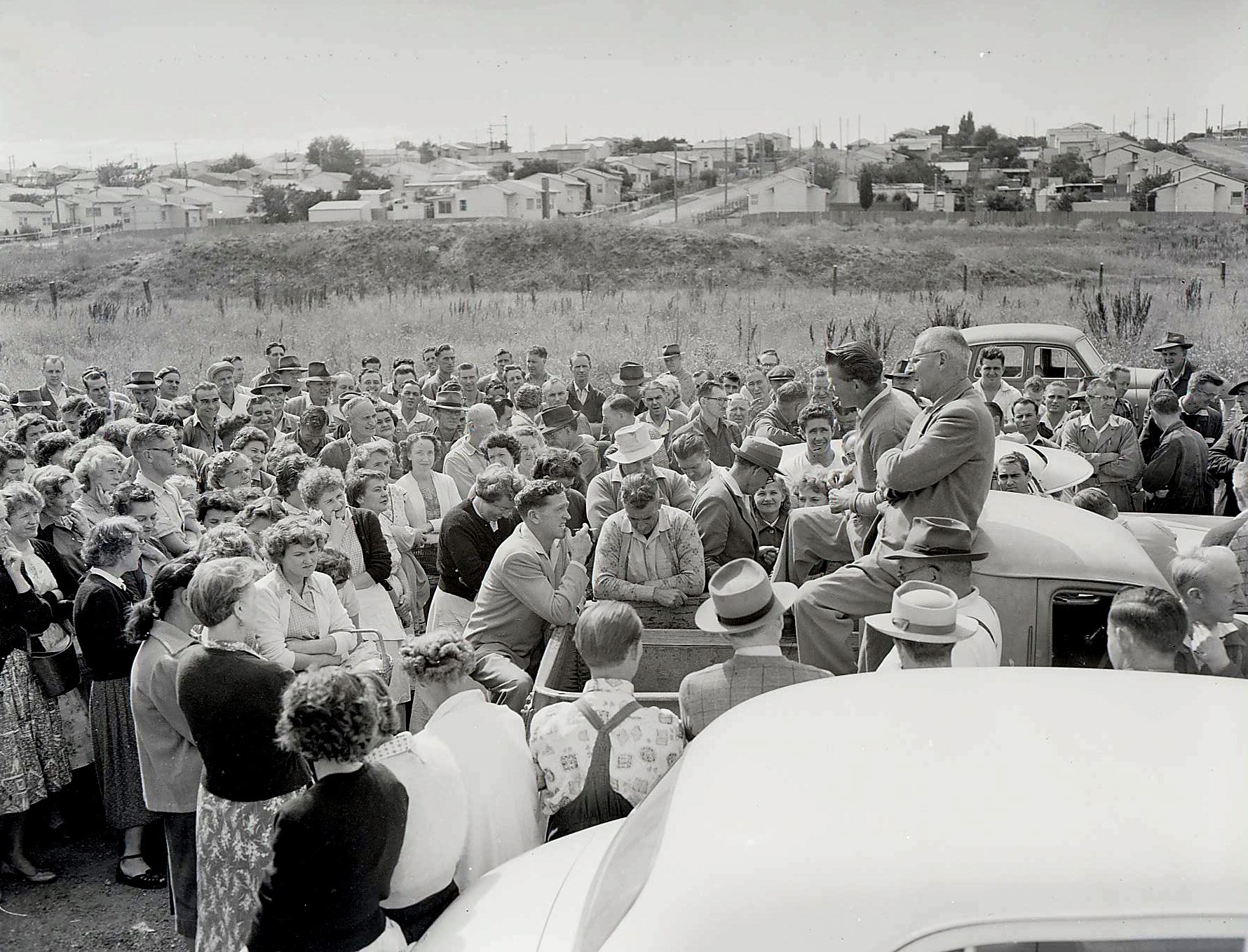 A black and white photograph of people in a paddock some standing, others sitting on the back of a truck