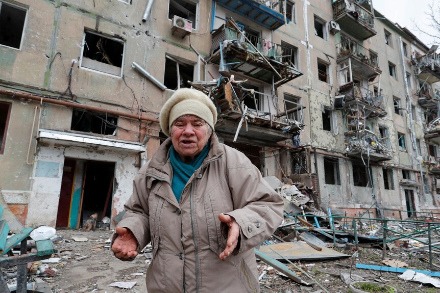 An elderly person wearing winter clothes stands and speaks outside of a badly damaged apartment building.