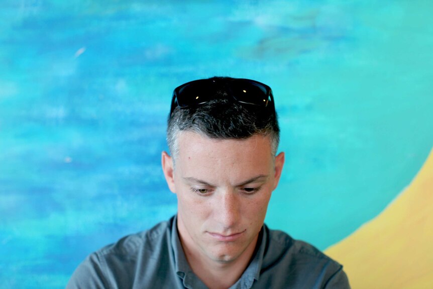 A man sits in front of a colourful wall looking down.