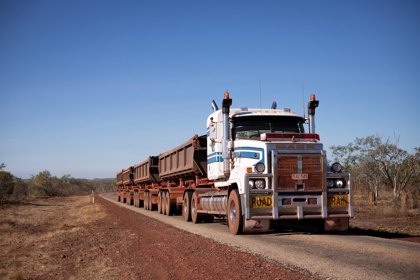 A road train with four trailers drives along a one-lane bitumen road. 