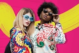 Two women pose with sunglasses and bright clothing against a pink wall for a story about how to accessorise.