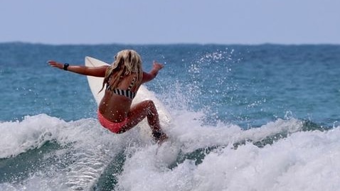 A tanned girl with long blonde hair surfing on a wave with blue ocean in the background on a sunny day