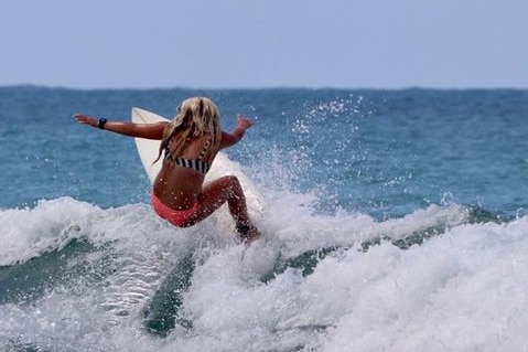 A tanned girl with long blonde hair surfing on a wave with blue ocean in the background on a sunny day