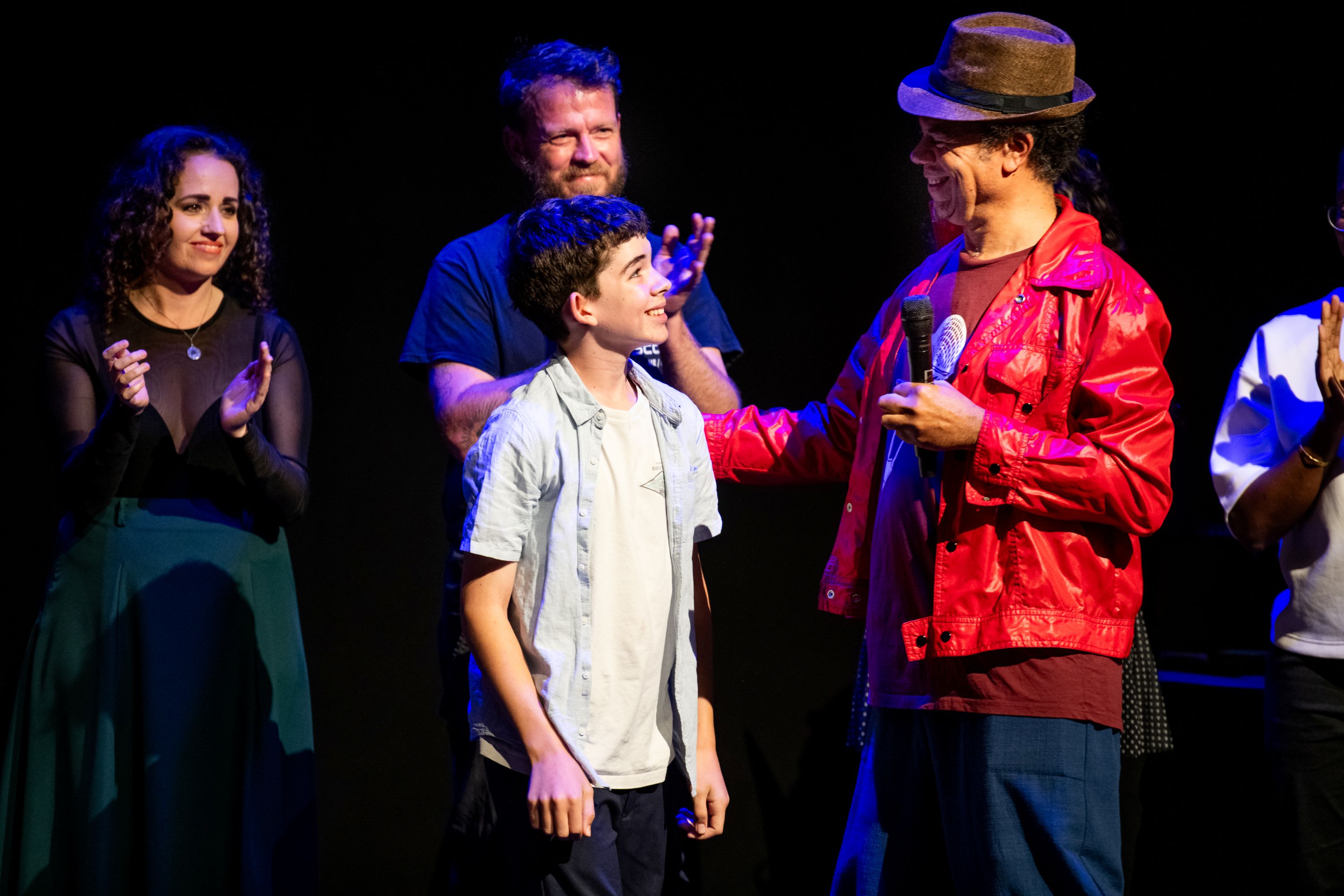 A boy looks up at a man wearing a hat and a red shirt who is holding a microphone, while two people in the background applaud.