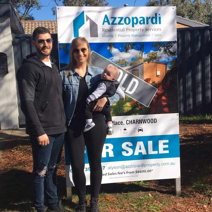A young family stand in front of a sold sign for their first home.