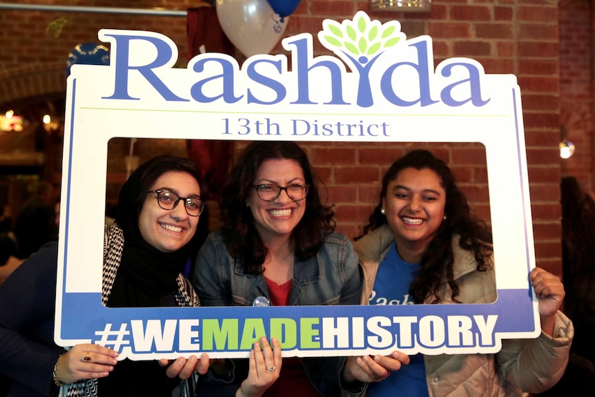 Three smiling women hold up a cardboard sign encircling their faces that says 'We Made History'.
