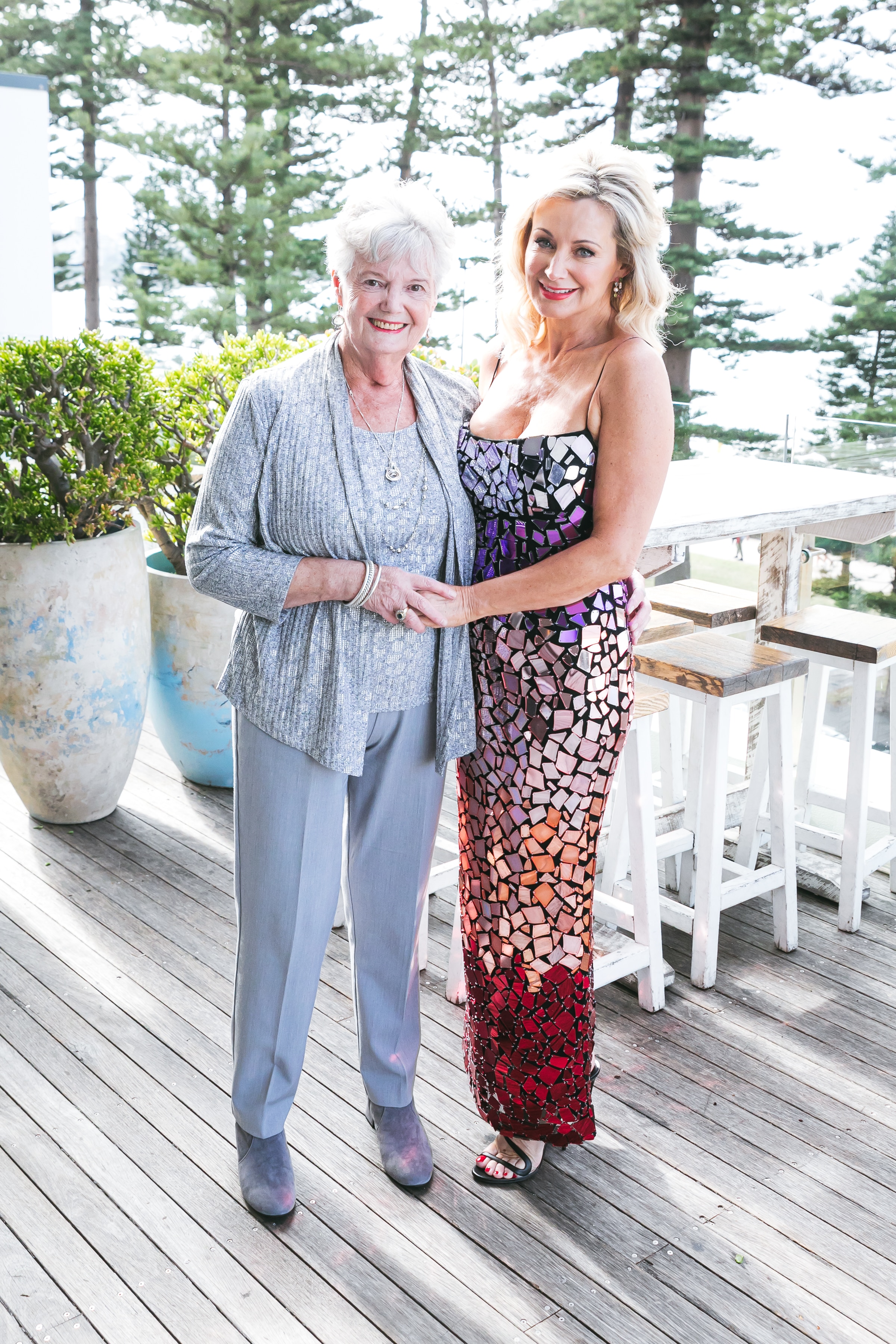 A woman and her daughter stand together, smiling while posing with pine trees in the background.