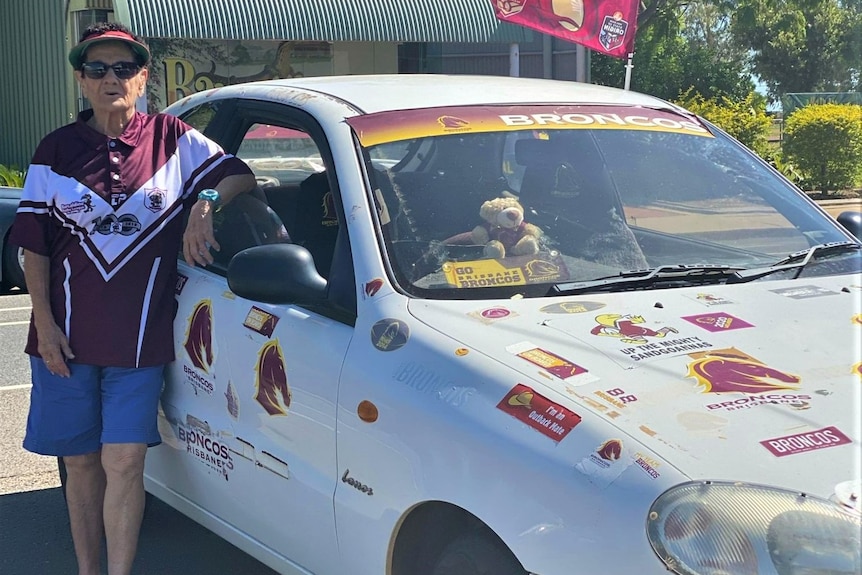 A lady in a cap and maroon jersey stands beside her white car white is covered in stickers of the broncos football team