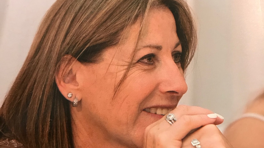 Middle-aged woman smiles and rests her chin on her hands. She wears diamond rings on her fingers and and diamond earrings