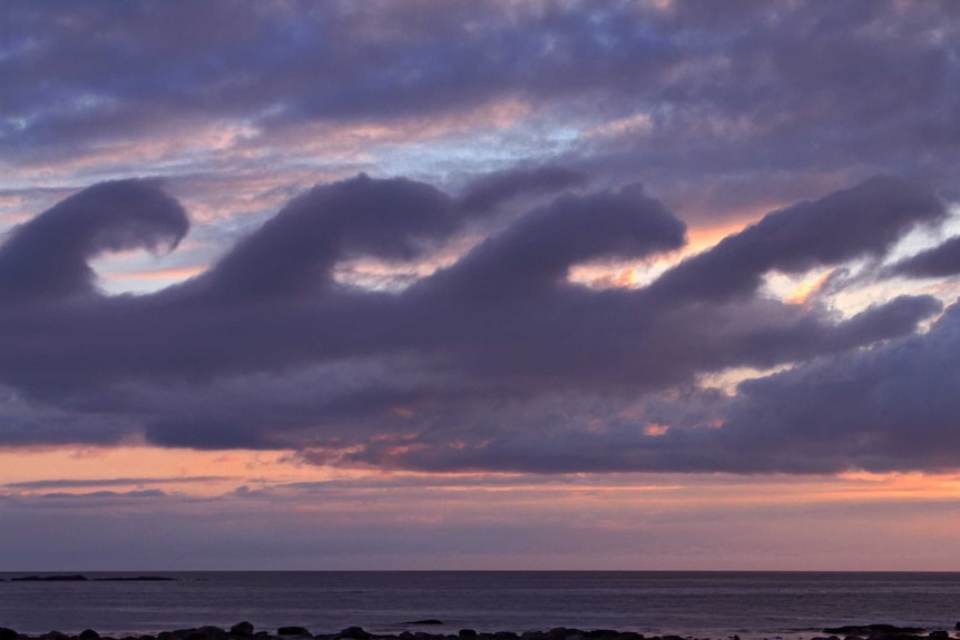 Four cloud waves break from left to right as sunrays colour the sky orange over a calm ocean