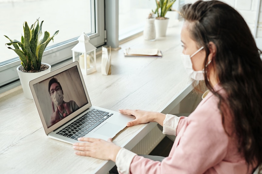 A woman with long dark hair and a face mask on joins a video call on her laptop