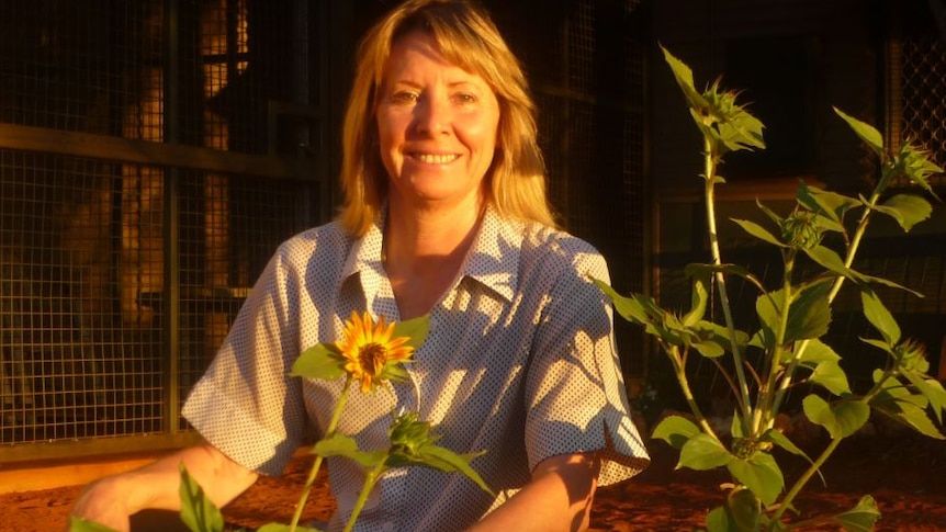 A women smiles among a group of sunflowers as the afternoon sun hits her face. She's in a front yard with red earth ground.