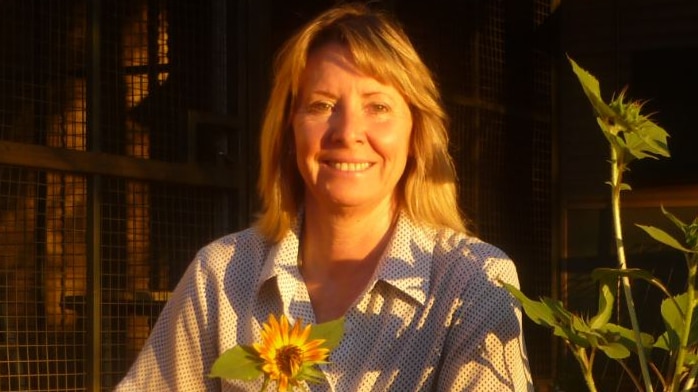 A women smiles among a group of sunflowers as the afternoon sun hits her face. She's in a front yard with red earth ground.