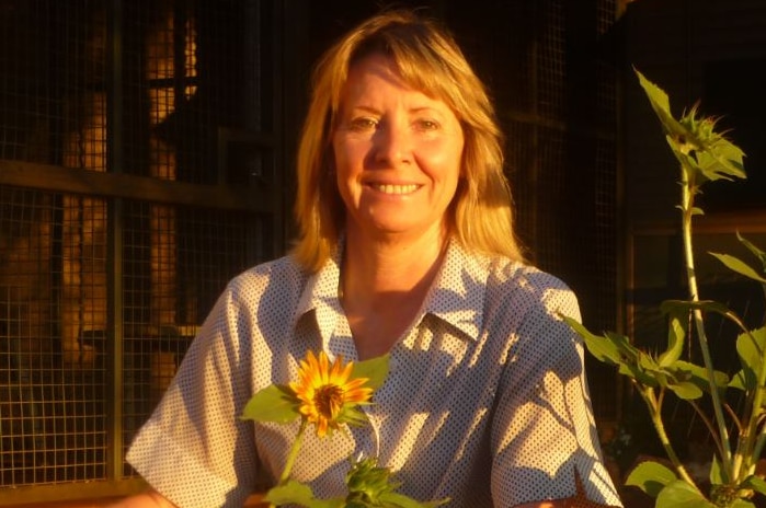 A women smiles among a group of sunflowers as the afternoon sun hits her face. She's in a front yard with red earth ground.