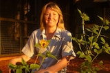 A women smiles among a group of sunflowers as the afternoon sun hits her face. She's in a front yard with red earth ground.