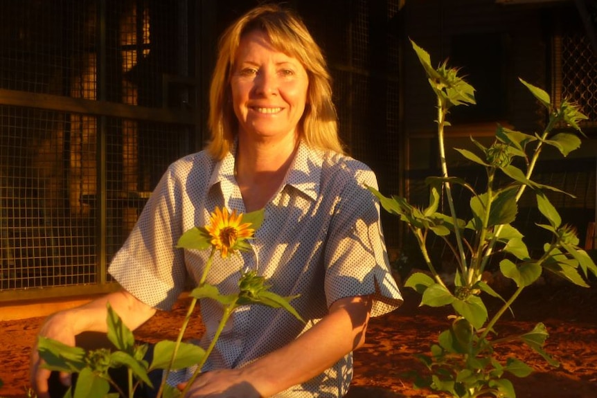 A women smiles among a group of sunflowers as the afternoon sun hits her face. She's in a front yard with red earth ground.
