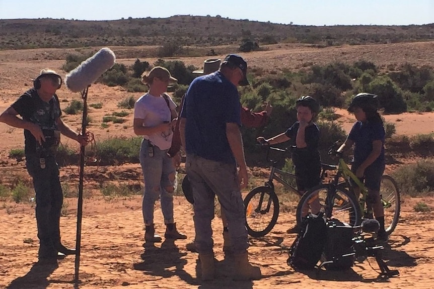 A group of people standing on dirt road, including one holding a boom microphone and two young children on bikes