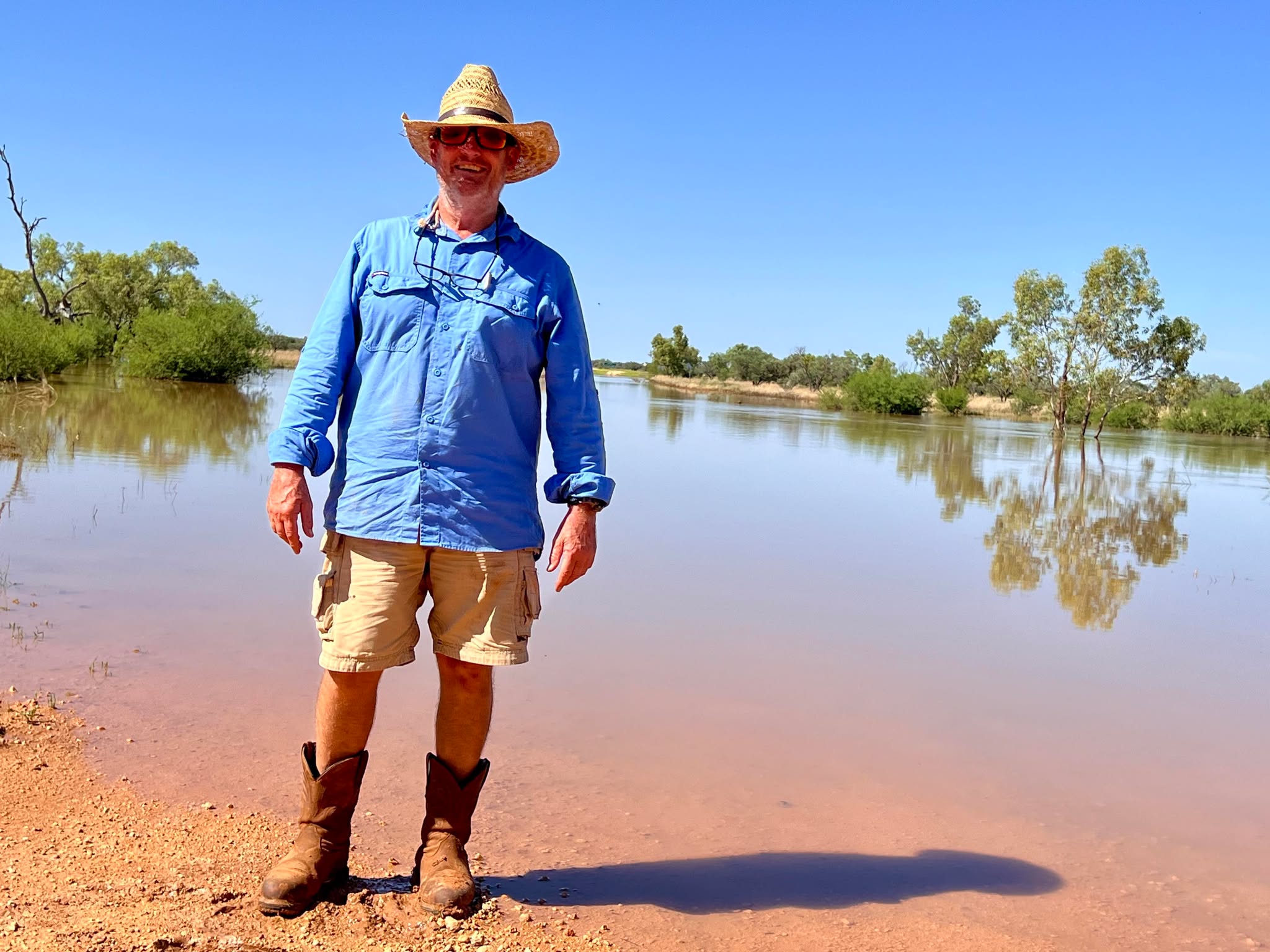 Man standing on red dirt, edge of water expanding well into distance behind him, trees appear in background