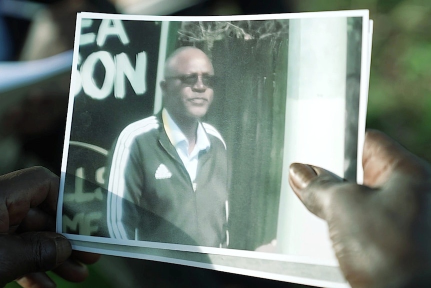 A person's hand holding a photograph of a man wearing a tracksuit top and sunglasses