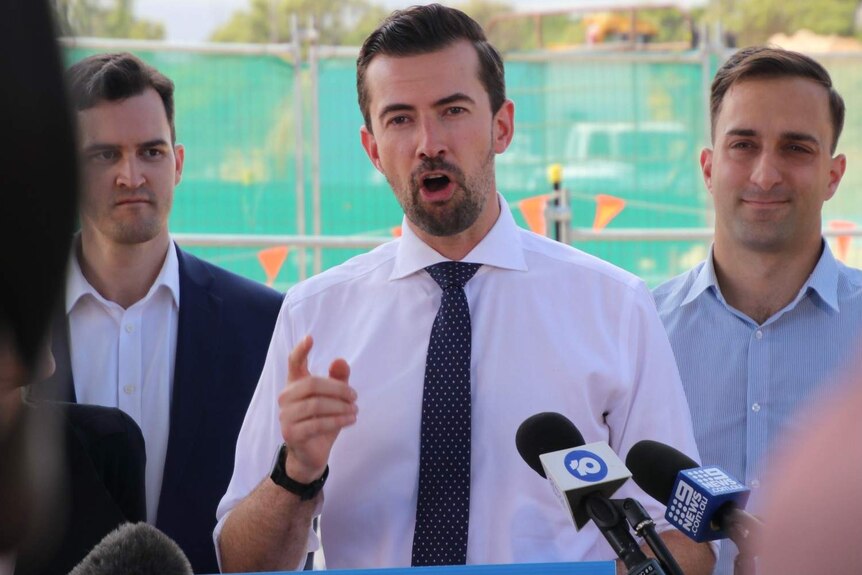 Zak Kirkup wearing a white shirt and blue tie, standing in front of two other men, at a podium for an election announcement.