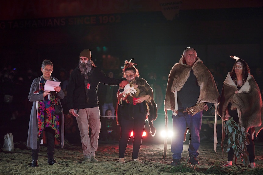 At night, five Aboriginal Tasmanians stand in a row some in possum skin cloaks in a welcome to country. Aunty Netty Shaw speaks