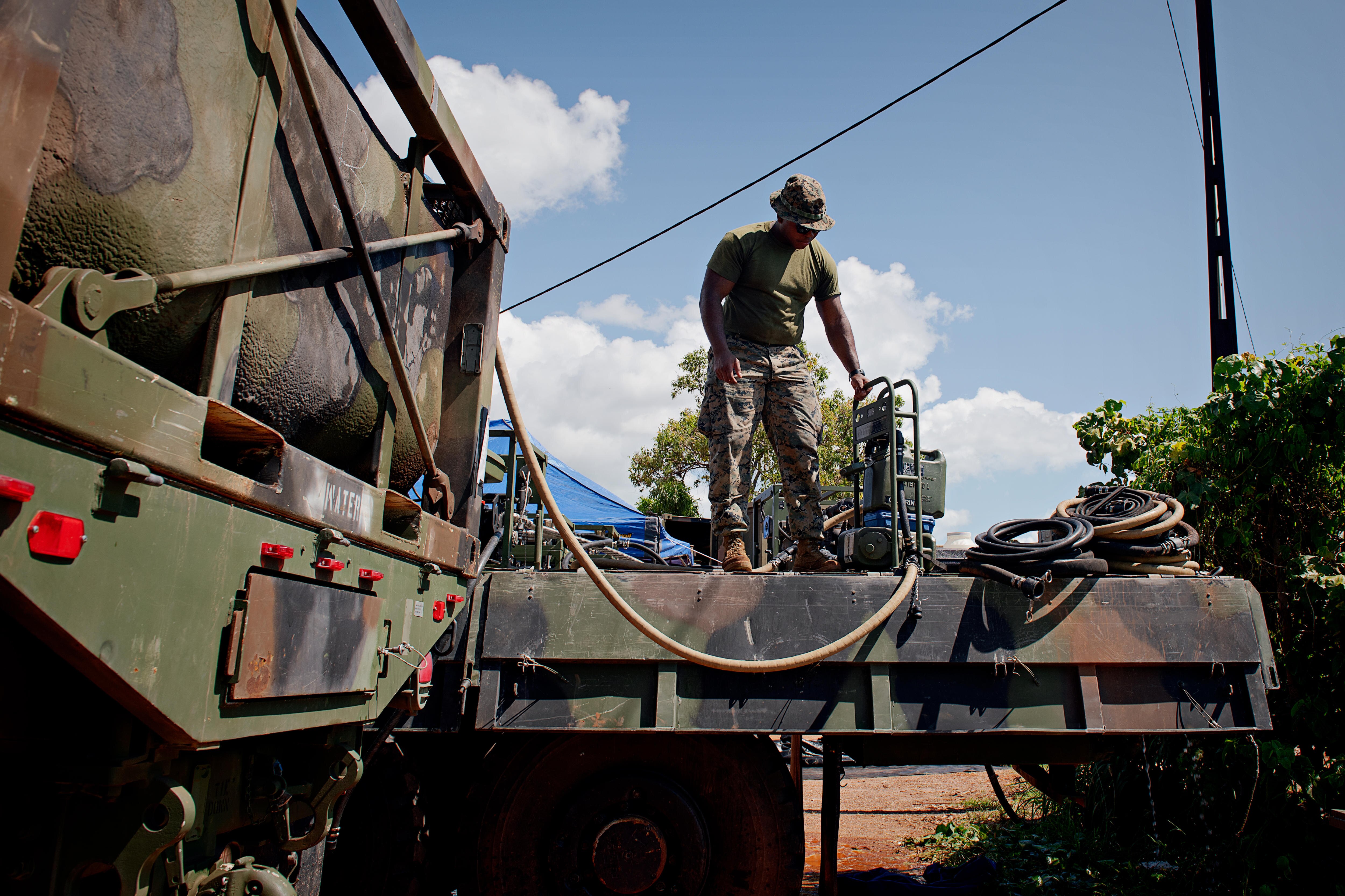 A US Marine in camo fatigues standing on the tray of an army truck, next to a small machine connected to a hose