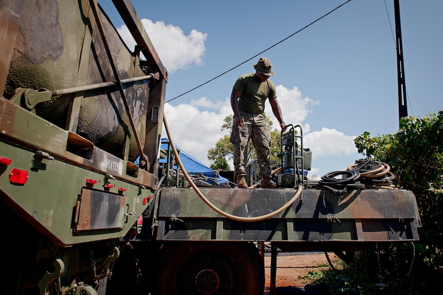 A US Marine in camo fatigues standing on the tray of an army truck, next to a small machine connected to a hose