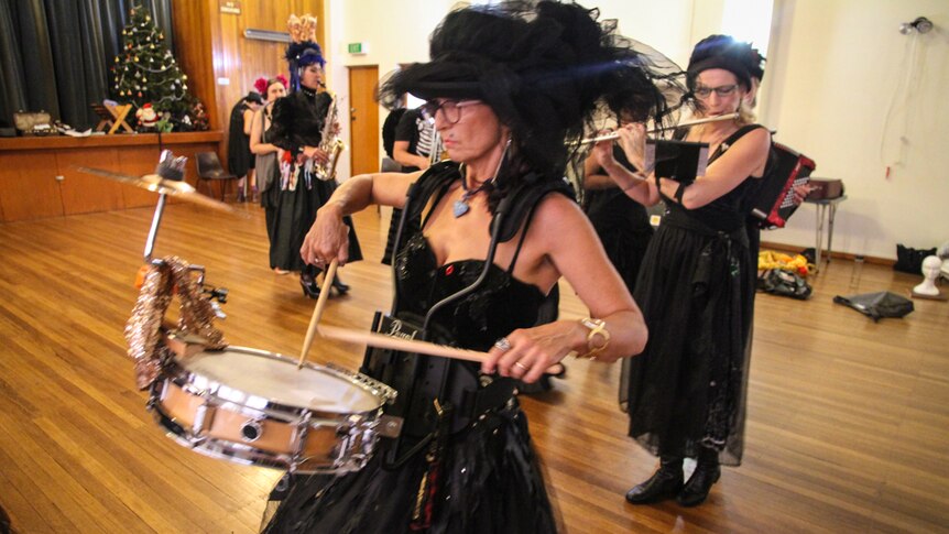A woman, dressed in black, plays the drums with other brass band performers in a church hall