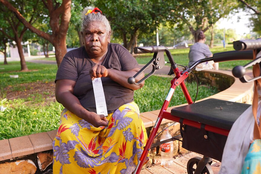 An Indigenous woman with a walker sitting outside in a park, holding a fine