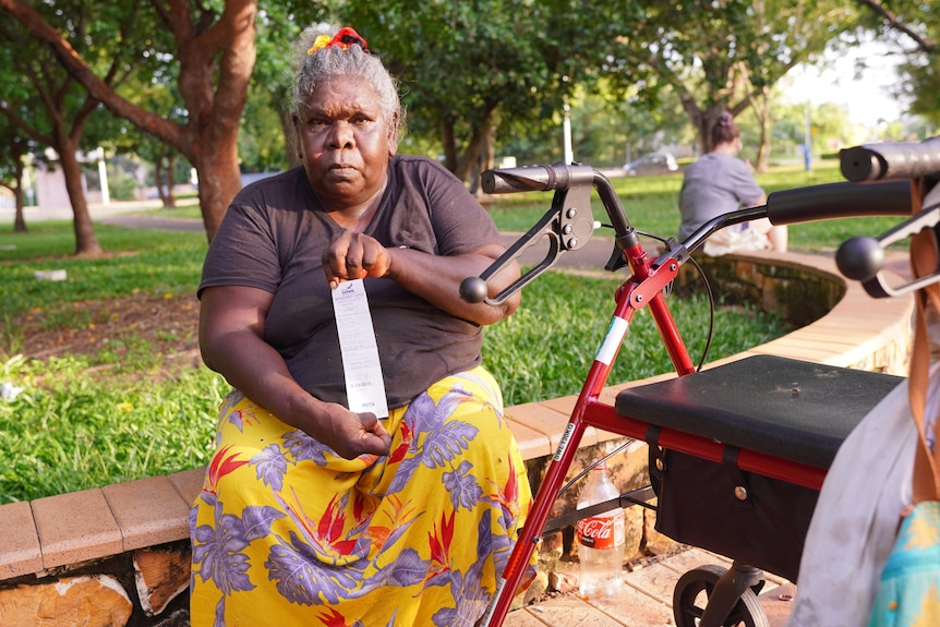 An Indigenous woman with a walker sitting outside in a park, holding a fine