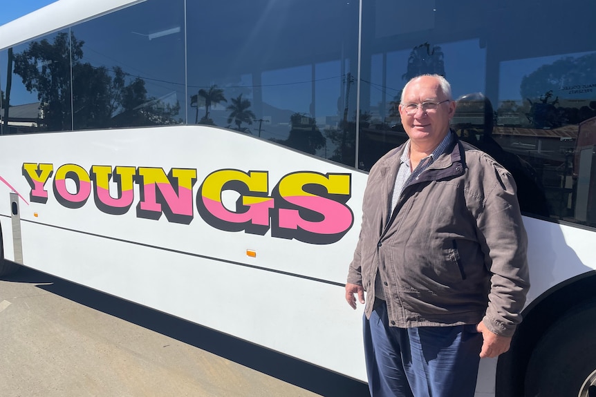 Man in brown jacket standing in front of bus with yellow and pink text reading Young's