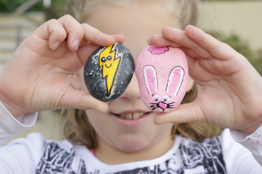 A young girl is holding up two painted rocks over her eyes.