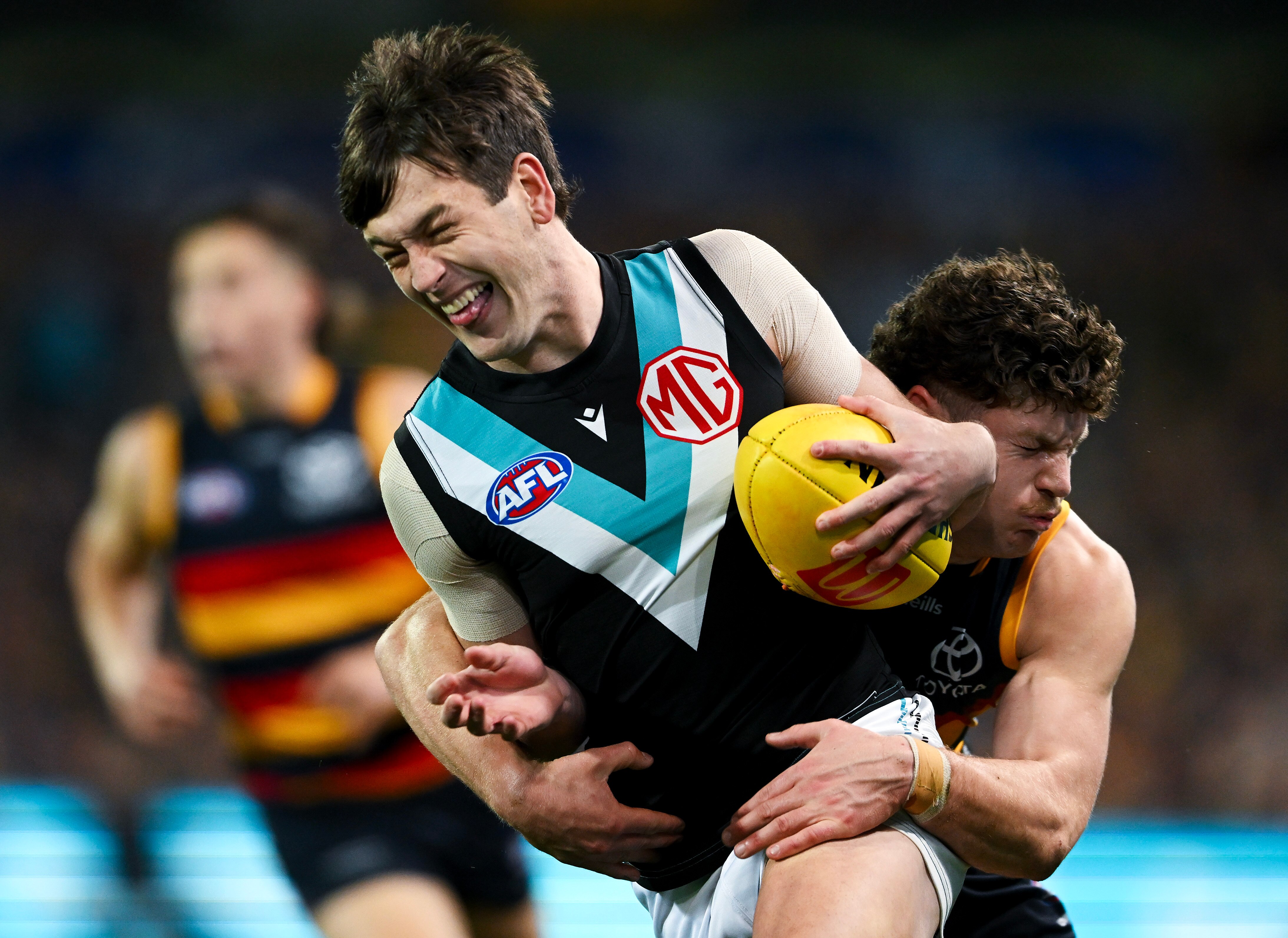 A Port Adelaide AFL player grimaces as he hangs onto the ball while being tackled from behind by a Crows defender.