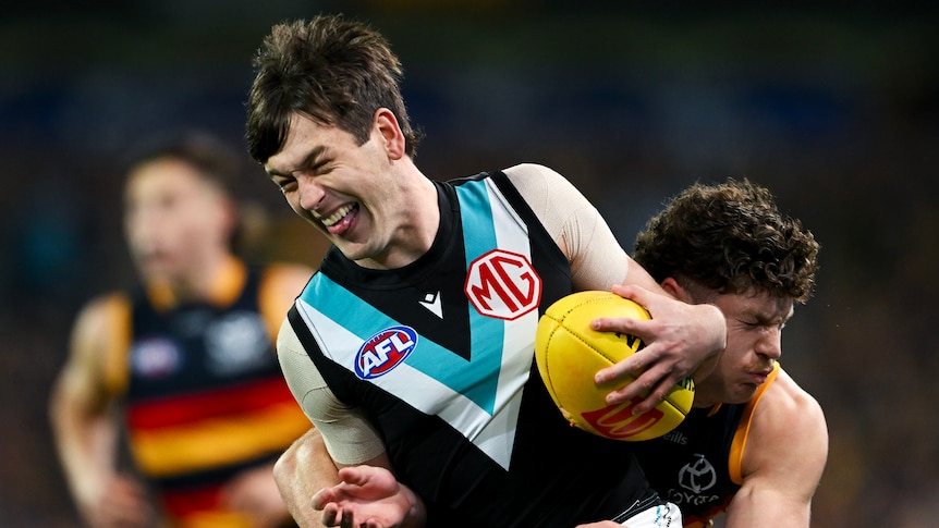 A Port Adelaide AFL player grimaces as he hangs onto the ball while being tackled from behind by a Crows defender.