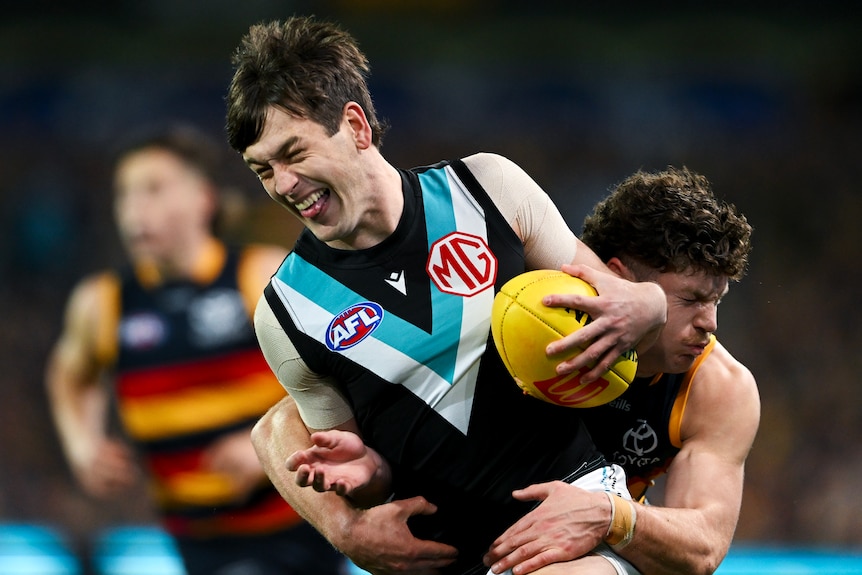 A Port Adelaide AFL player grimaces as he hangs onto the ball while being tackled from behind by a Crows defender.