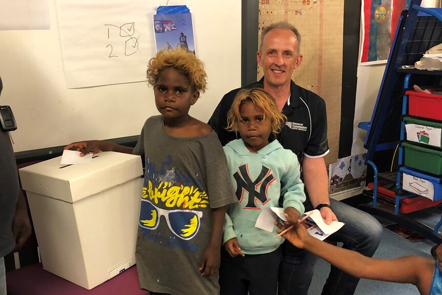 A man poses with two young students as they use a voting booth