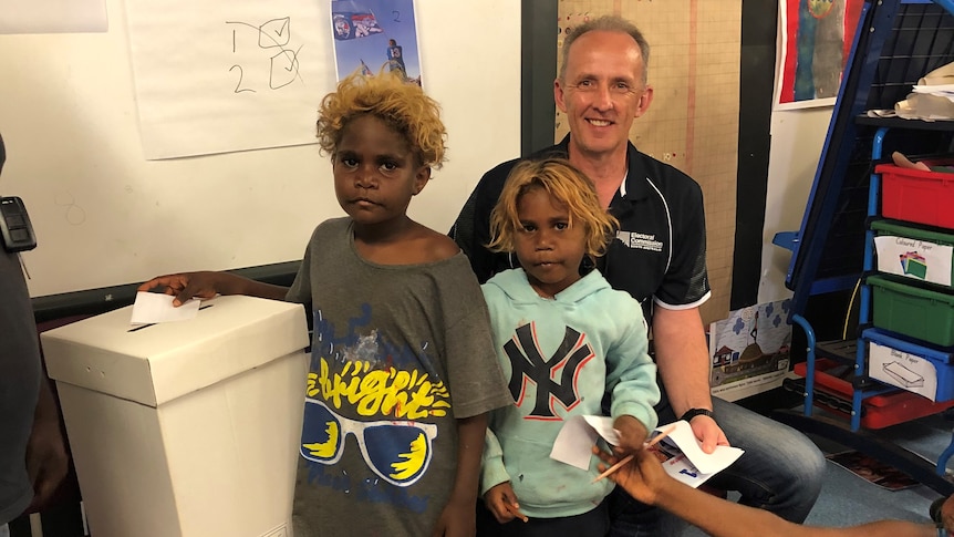 A man poses with two young students as they use a voting booth