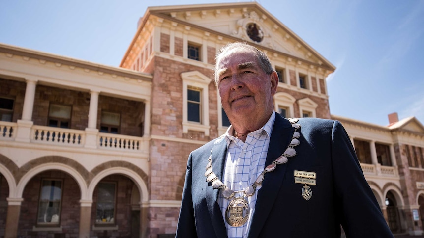 An older man in a blazer wears a municipal medallion and stands in front of a historic-looking building.