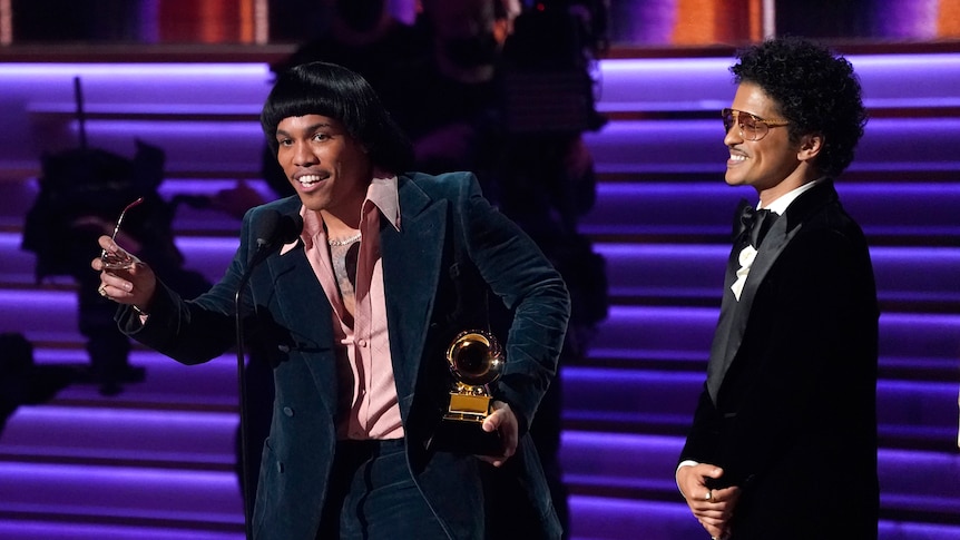 anderson .paak speaks into a microphone on stage holding sunglasses and a grammy. bruno mars smiles next to him