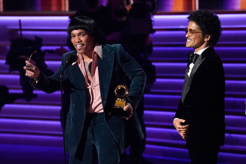 anderson .paak speaks into a microphone on stage holding sunglasses and a grammy. bruno mars smiles next to him