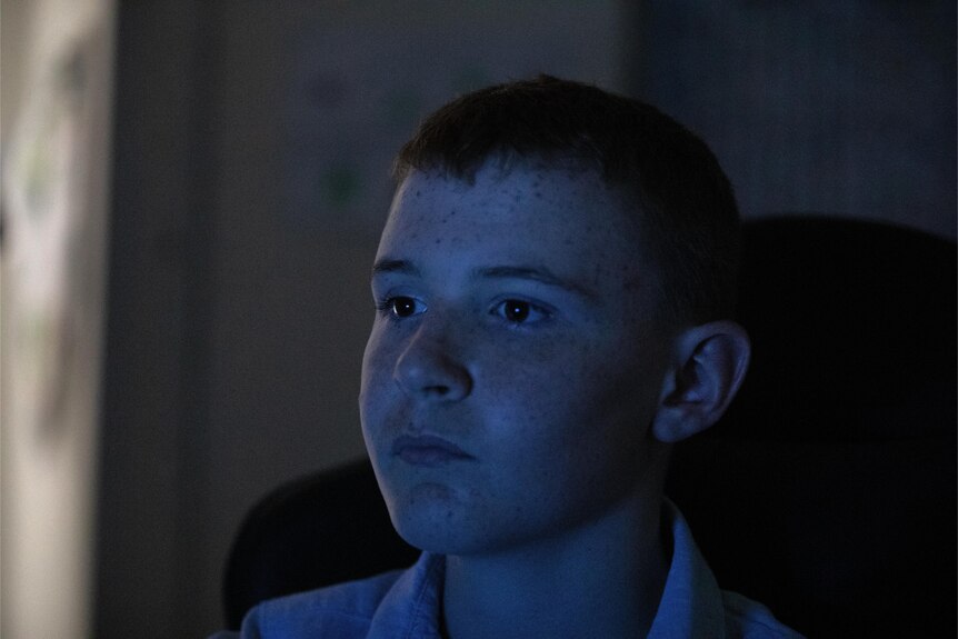 A close up of a teenage boy's face as he sits in a dark room looking at a screen with blue light reflecting on his face 