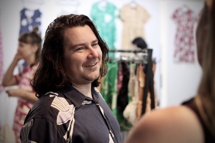 Troy Casey smiles, with colourful clothing hanging behind him.