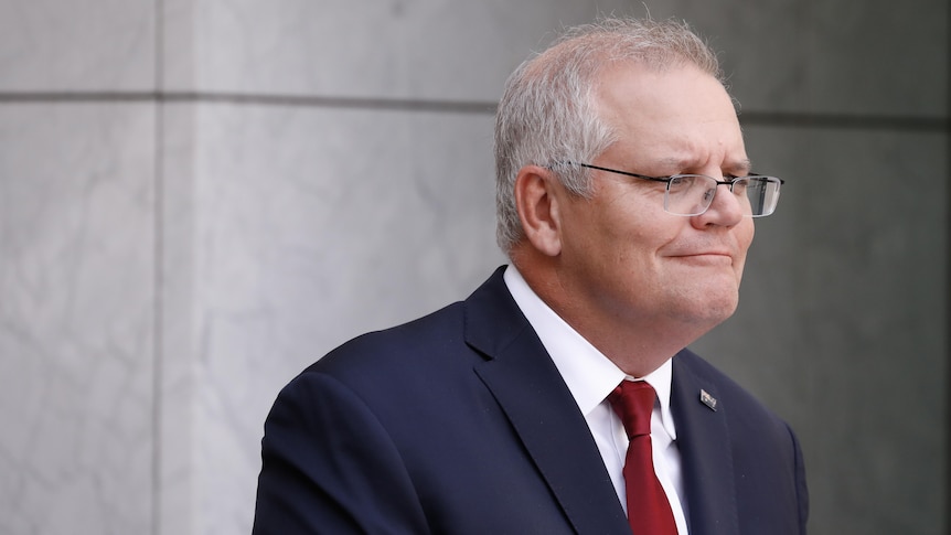 Morrison is wearing a dark blue suit and red tie, with a slight grin.
