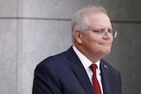 Morrison is wearing a dark blue suit and red tie, with a slight grin.