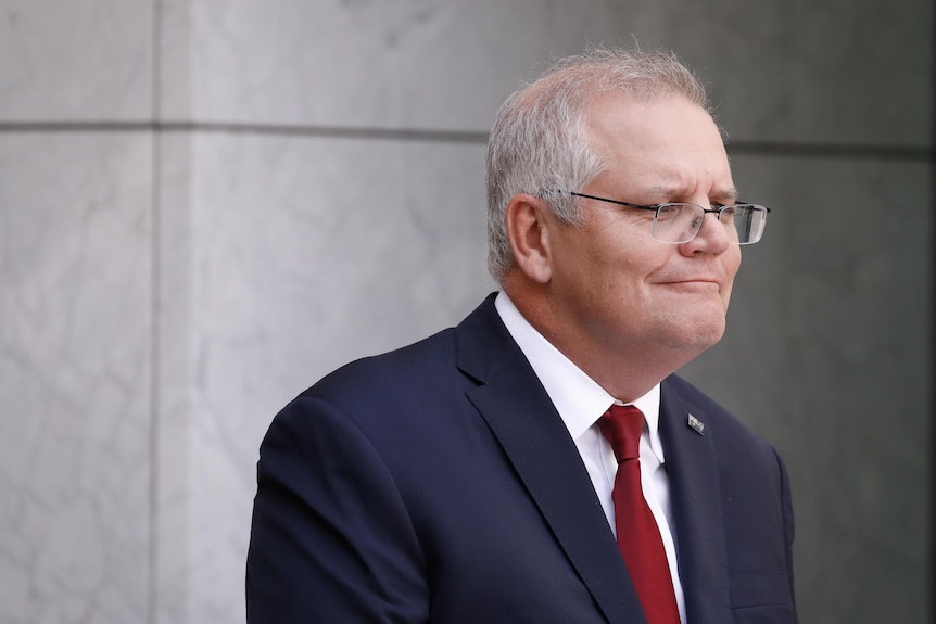 Morrison is wearing a dark blue suit and red tie, with a slight grin.