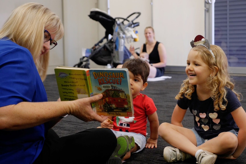 A woman holding a picture book reads to a room, two children are sitting in front of her looking at the book. 