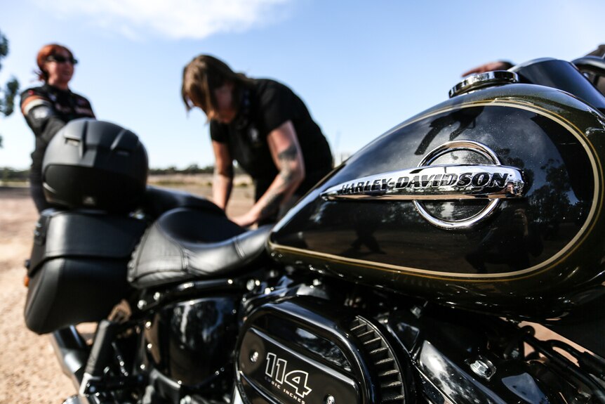 A close up of a Harley-Davidson, with some women, out of focus, standing behind it.