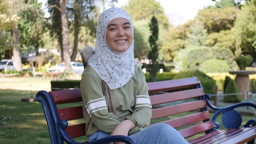 Young woman wearing a hijab sits on park bench smiling