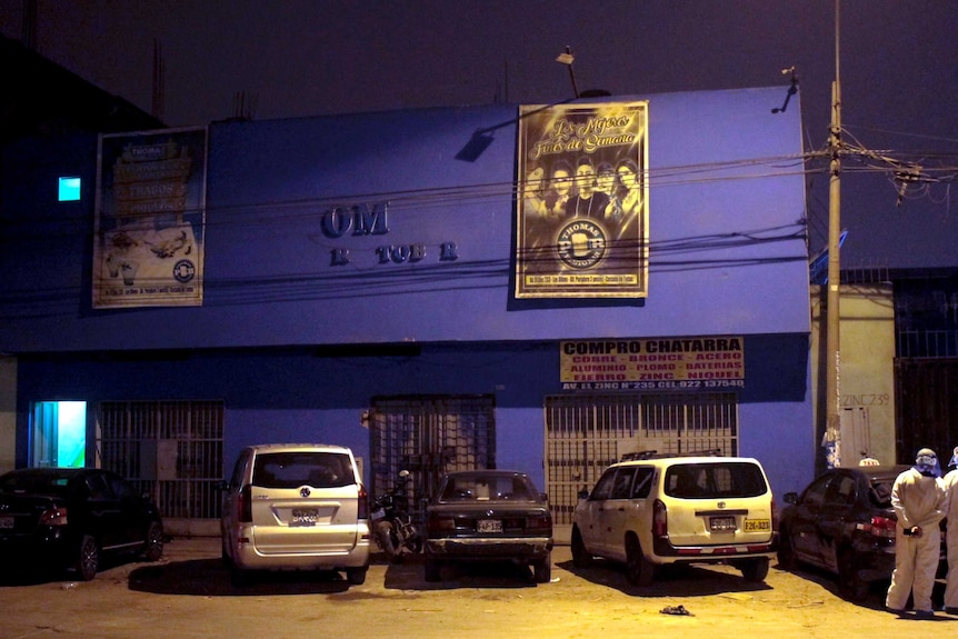 Police officers wearing protective clothing as they stand in the night outside in the carpark of a blue building.